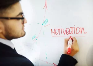 Homme portant des lunettes écrivant le mot motivation en rouge sur un tableau blanc à côté d'un graphique de progression.